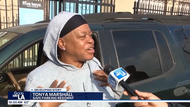 A woman in a gray hoodie and black headscarf speaks to a news reporter holding a Good Day LA microphone outside near a black vehicle. A caption identifies her as Tonya Marshall, Safe Parking resident.
