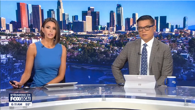 Two news anchors sit at a desk in a studio with a city skyline backdrop. The screen displays 