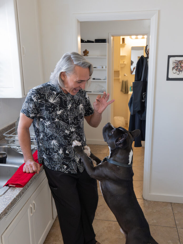 A man standing in a kitchen smiles and gives a hand signal to a black and white dog with its front paws on his leg.