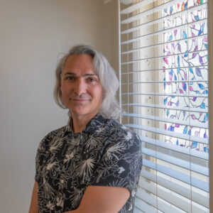 A person with shoulder-length gray hair and a patterned shirt stands by a window with white blinds and colorful window film in a softly lit room.