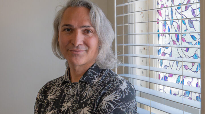 A person with shoulder-length gray hair and a patterned shirt stands by a window with white blinds and colorful window film in a softly lit room.