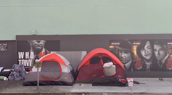 Two tents and personal belongings are set up on a city sidewalk in front of a mural featuring black-and-white movie character faces.