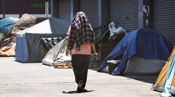 A person walks past tents on a sidewalk, covering their head with a plaid blanket. The area appears to be a homeless encampment with closed storefronts in the background.
