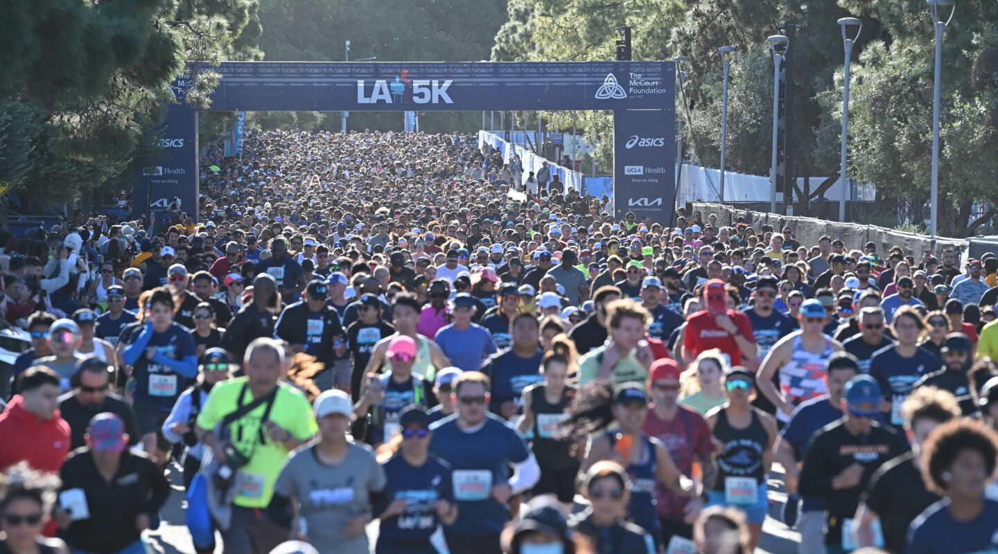 Large crowd of runners participates in a 5K race, moving along a tree-lined path under a banner that reads “LA 5K.” Most runners wear race bibs and athletic attire.