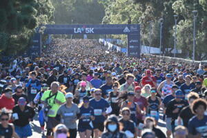 Large crowd of runners participates in a 5K race, moving along a tree-lined path under a banner that reads “LA 5K.” Most runners wear race bibs and athletic attire.